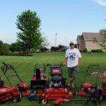 Man standing in a green yard posing for a photo surrounded by different models of Toro lawnmowers