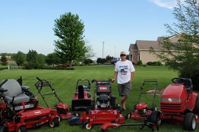 Man standing in a green yard posing for a photo surrounded by different models of Toro lawnmowers