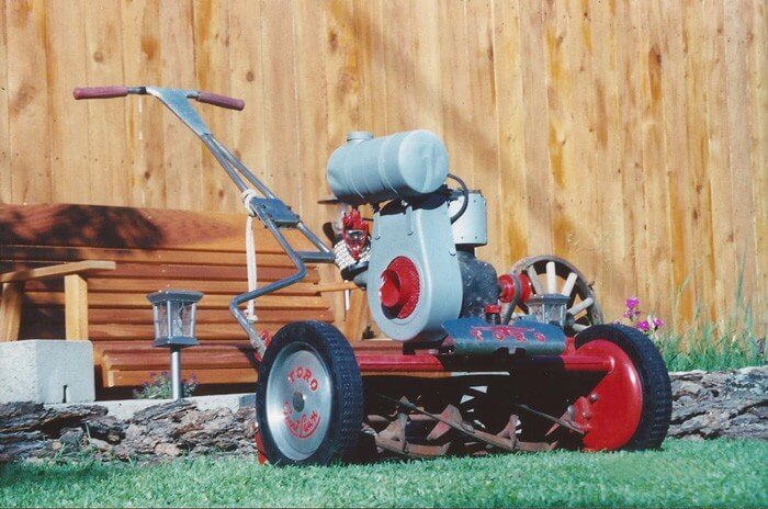 Image of older model of Toro lawnmower in a yard with a brown bench in the background