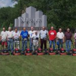 Group of 12 men posing for a photo in front of the Minot city sign each with the same model of lawnmower.