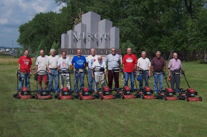 Group of 12 men posing for a photo in front of the Minot city sign each with the same model of lawnmower.