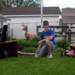 Man kneeling in lush green grass posing for a photo with a lawn mower on either side of him