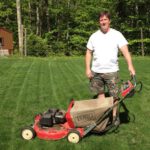 A man posing with his Toro lawnmower standing in green freshly mowed grass featuring trees and a brown shed in the back