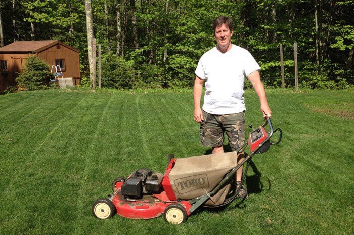 A man posing with his Toro lawnmower standing in green freshly mowed grass featuring trees and a brown shed in the back