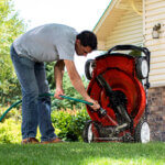 A man leaning over to clean the bottom of his lawnmower with a hose