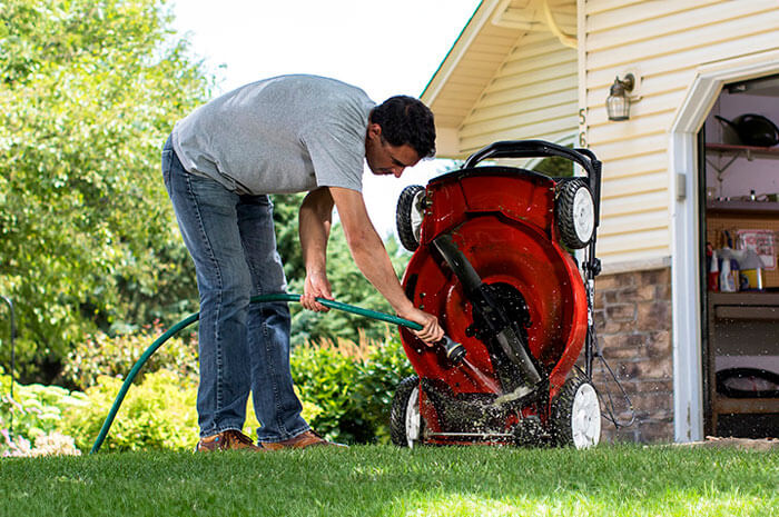 A man leaning over to clean the bottom of his lawnmower with a hose