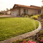 A sprinkler watering green grass outside of a brown home