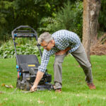 Man standing in front of his lawnmower, leaning over to clear brush out of the mowers path