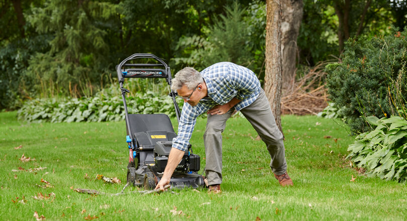 Man standing in front of his lawnmower, leaning over to clear brush out of the mowers path
