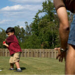 Child about to hit a T Ball with his dad in their backyard