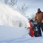 Man in brown jacket using snowblower to clear snow from sidewalk