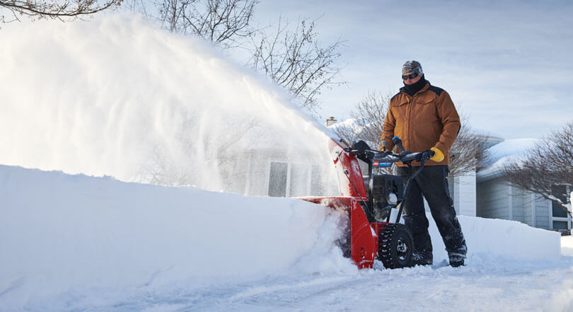 Man in brown jacket using snowblower to clear snow from sidewalk