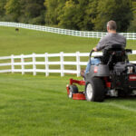 Man using Toro riding mower in green yard surrounded by white fence with horses in the background