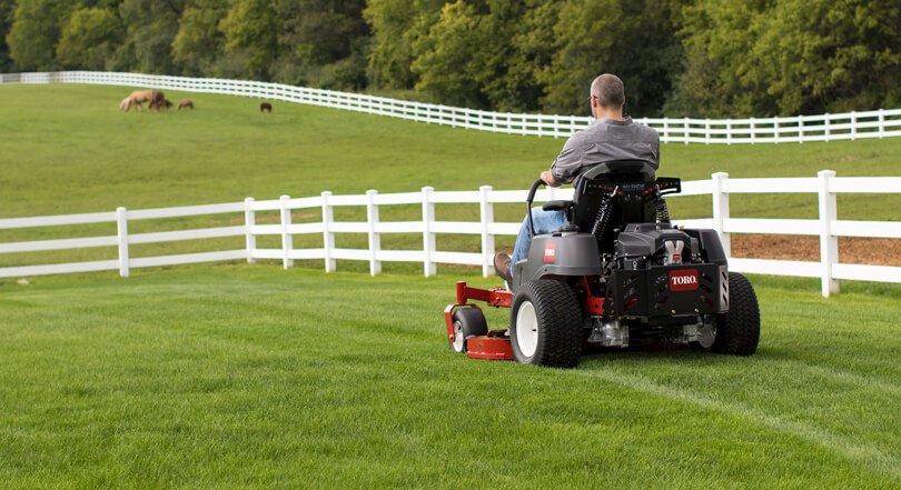 Man using Toro riding mower in green yard surrounded by white fence with horses in the background